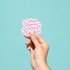 May include: A white sticker with pink lettering that reads "Very Demine, Very Much!" with pink flower accents.