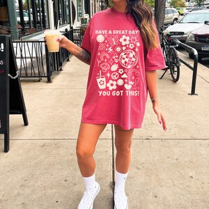 Puede incluir: Una camiseta roja de gran tama&ntilde;o con un estampado gr&aacute;fico blanco de varios iconos y el texto "Have a Great Day!" y "You Got This!"