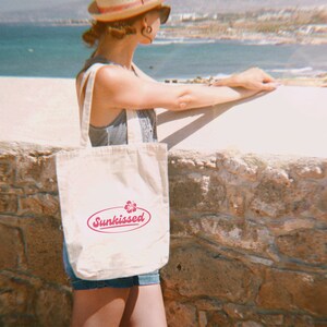May include: A white canvas tote bag with a pink logo that says "Sunkissed" and a pink flower graphic. The bag is being held by a person wearing a straw hat and sunglasses.