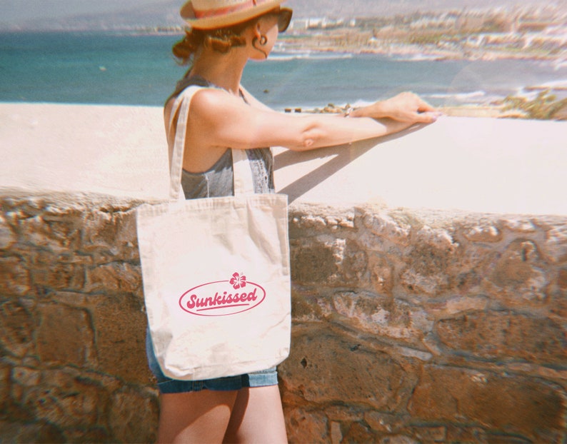 May include: A white canvas tote bag with a pink logo that says "Sunkissed" and a pink flower graphic. The bag is being held by a person wearing a straw hat and sunglasses.