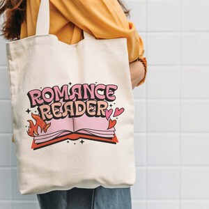 May include: A white canvas tote bag with a pink and red graphic design. The design features a book with flames and the text "Romance Reader".