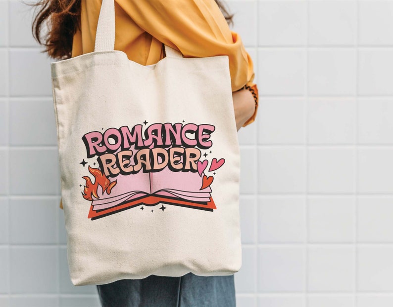 May include: A white canvas tote bag with a pink and red graphic design. The design features a book with flames and the text "Romance Reader".