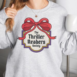 May include: A light grey sweatshirt with a graphic of a red bow and the text "The Thriller Readers Society" in a retro style. The design is framed by a purple and gold border. A white mug is held in the hand.