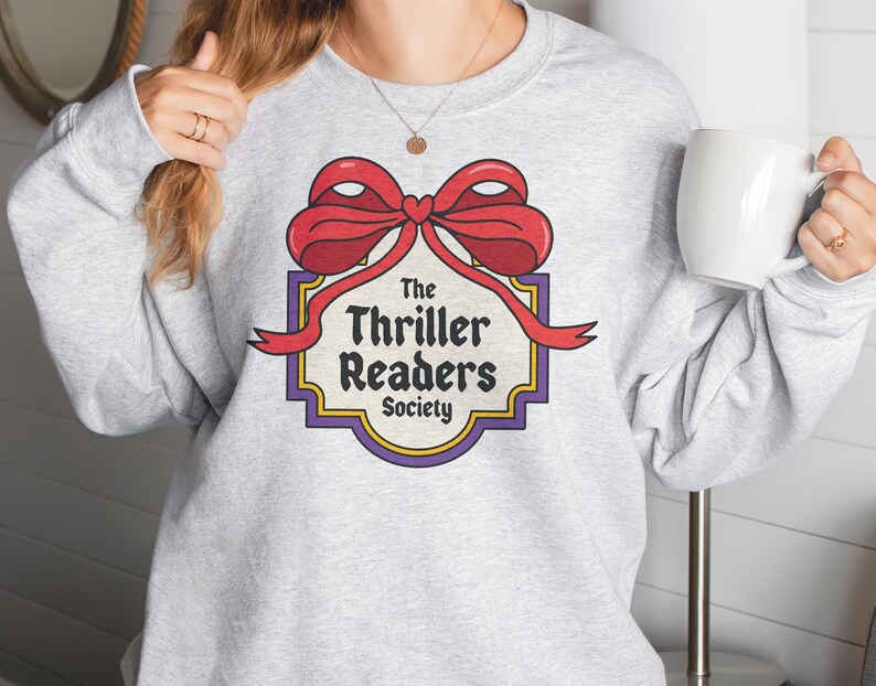 May include: A light grey sweatshirt with a graphic of a red bow and the text "The Thriller Readers Society" in a retro style. The design is framed by a purple and gold border. A white mug is held in the hand.