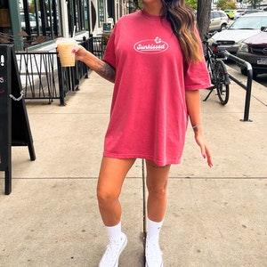 May include: A woman wearing a red oversized t-shirt with the text "Sunkissed" in a white oval. She is holding a cup of coffee and wearing white sneakers.