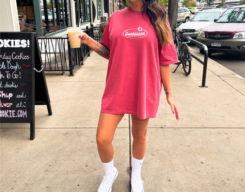 May include: A woman wearing a red oversized t-shirt with the text "Sunkissed" in a white oval. She is holding a cup of coffee and wearing white sneakers.