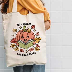 May include: A white canvas tote bag with a cute illustration of a smiling pumpkin reading a book. The text on the bag reads "Fall Breeze and Cozy Reads".