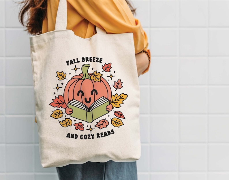 May include: A white canvas tote bag with a cute illustration of a smiling pumpkin reading a book. The text on the bag reads "Fall Breeze and Cozy Reads".