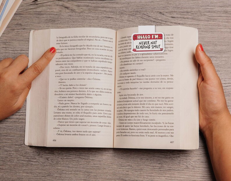 May include: A close-up of a book with a bookmark that reads "Hello I'm Never Not Reading Smut". The bookmark is white with red and black text. The book is open to a page with Spanish text.