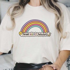 May include: White t-shirt with a rainbow and the text "Think Happy Thoughts" printed on it.