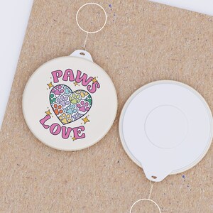 May include: A white round button with a pink heart design and the text "PAWS LOVE" in black. The heart is filled with paw prints in various colors. The button is attached to a silver key ring.