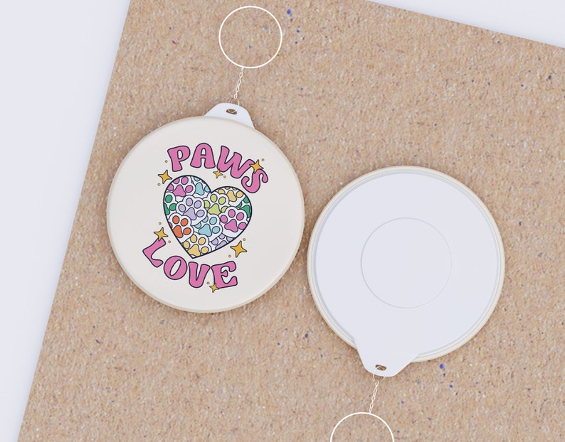 May include: A white round button with a pink heart design and the text "PAWS LOVE" in black. The heart is filled with paw prints in various colors. The button is attached to a silver key ring.