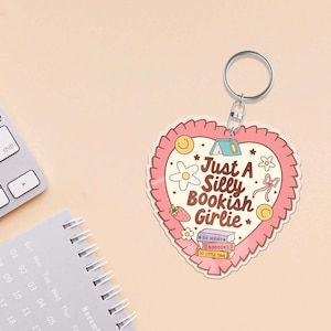 May include: A heart-shaped keychain with a pink ruffled border and the text "Just A Silly Bookish Girlie." The design includes illustrations of a tent, flowers, a strawberry, and books. A silver key ring is attached.