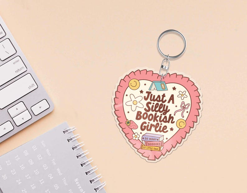 May include: A heart-shaped keychain with a pink ruffled border and the text "Just A Silly Bookish Girlie." The design includes illustrations of a tent, flowers, a strawberry, and books. A silver key ring is attached.