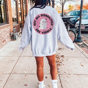 Op de afbeelding: Een grijs sweatshirt met een roze cirkelvormige afbeelding op de rug. De afbeelding toont een witte geest met een roze halo en de tekst "Not all disabilities are visible."