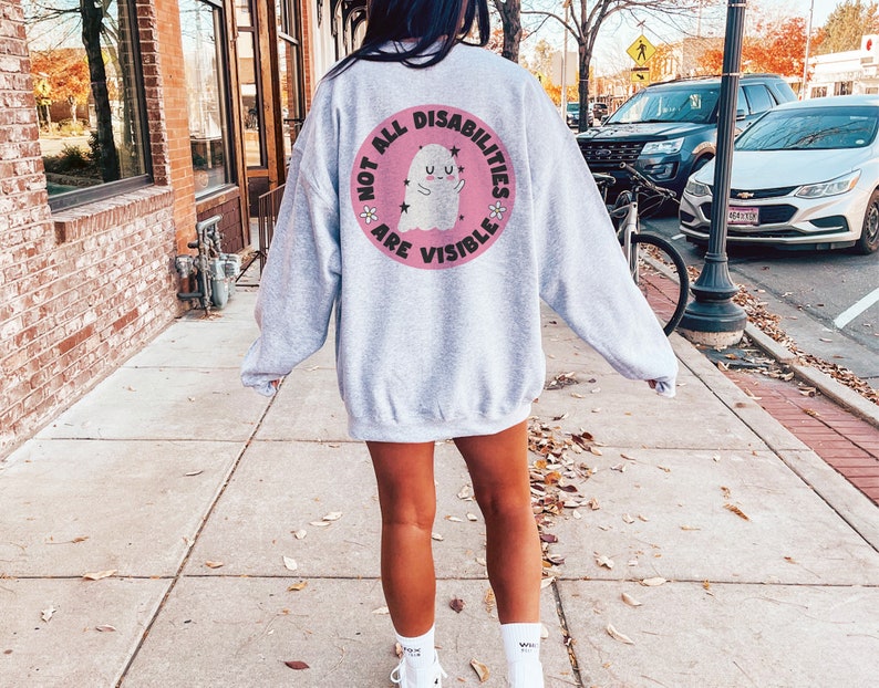 Op de afbeelding: Een grijs sweatshirt met een roze cirkelvormige afbeelding op de rug. De afbeelding toont een witte geest met een roze halo en de tekst "Not all disabilities are visible."