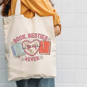 May include: A white canvas tote bag with the text "Book Besties 4ever" and a pink heart with a bow and two books on either side. The books are blue and red.