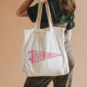 May include: A white canvas tote bag with pink text that reads "Tired AF" in a pennant shape.