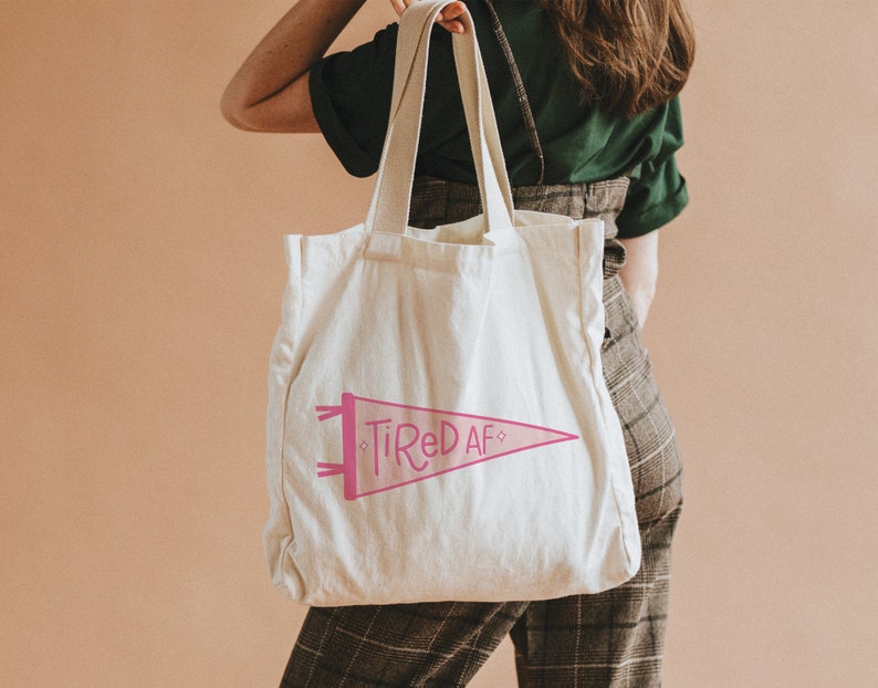 May include: A white canvas tote bag with pink text that reads "Tired AF" in a pennant shape.