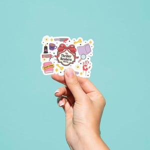 May include: A sticker with a collection of thriller-themed illustrations, including a knife, book, magnifying glass, and a handprint. The sticker has the text "The Thriller Readers Society" and is held against a teal background.