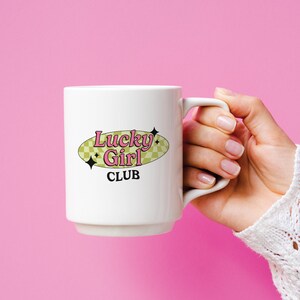 May include: White ceramic mug with a black and pink logo that says "Lucky Girl Club" on a yellow oval with a checkered pattern.