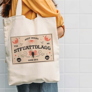 Puede incluir: Una bolsa de lona blanca con un dise&ntilde;o gr&aacute;fico rojo y negro. El gr&aacute;fico presenta un sol, una luna, estrellas, calaveras y el texto "SMUT BOARD", "YES", "NO", "STFUATTTLDRAGG" y "GOOD BYE".