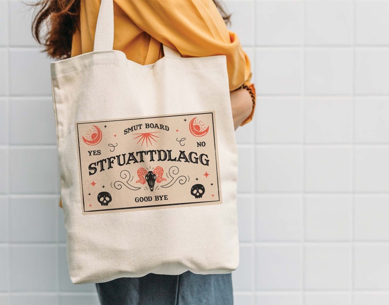 Puede incluir: Una bolsa de lona blanca con un dise&ntilde;o gr&aacute;fico rojo y negro. El gr&aacute;fico presenta un sol, una luna, estrellas, calaveras y el texto "SMUT BOARD", "YES", "NO", "STFUATTTLDRAGG" y "GOOD BYE".