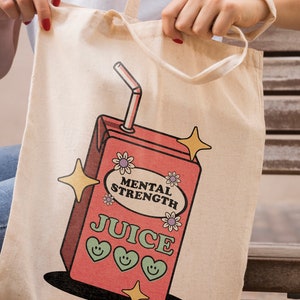 Puede incluir: Una bolsa de lona blanca con un dise&ntilde;o de caja de jugo de dibujos animados rosa y roja. La caja de jugo dice "Mental Strength Juice" y tiene tres corazones sonrientes en ella. Tambi&eacute;n hay estrellas brillantes y una pajita que sale de la parte superior.