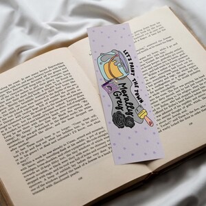 May include: A purple and white bookmark with a paint can, paintbrush, and flowers. The text on the bookmark reads "Let's paint the town Morally Grey".