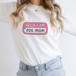 Peut inclure: Sweat-shirt blanc &agrave; manches longues avec une &eacute;tiquette de nom rose qui dit "Hello I am Dog Mom" avec des fleurs color&eacute;es et un smiley.
