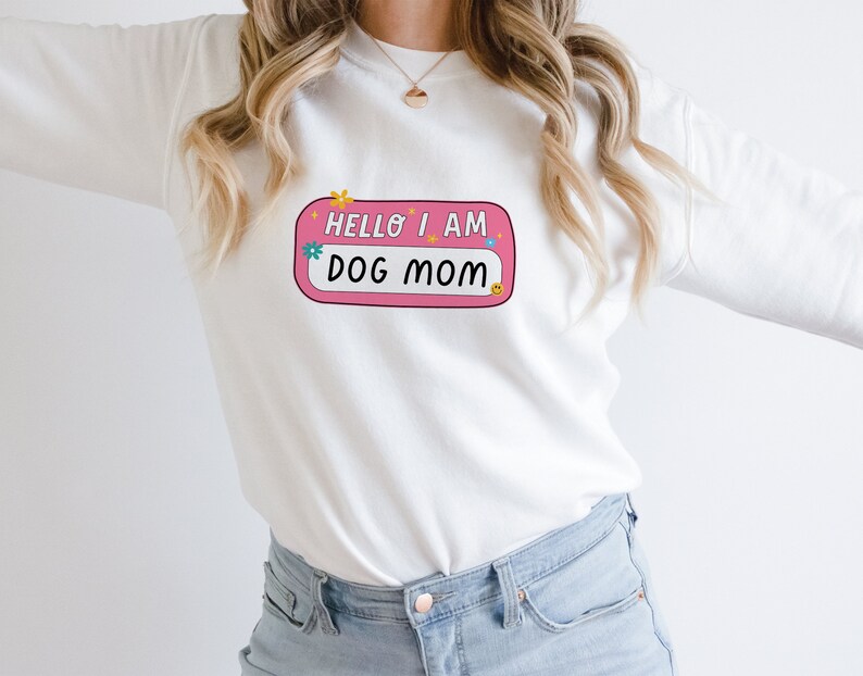 Peut inclure: Sweat-shirt blanc &agrave; manches longues avec une &eacute;tiquette de nom rose qui dit "Hello I am Dog Mom" avec des fleurs color&eacute;es et un smiley.