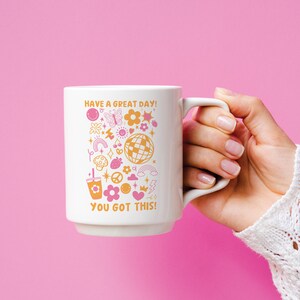 Puede incluir: Taza de cer&aacute;mica blanca con un dise&ntilde;o rosa y naranja que presenta una variedad de ilustraciones, incluyendo una mariposa, un arco&iacute;ris, un signo de paz, un coraz&oacute;n, una bola de discoteca y una cereza. El texto en la taza dice "Have a great day! You got this!"