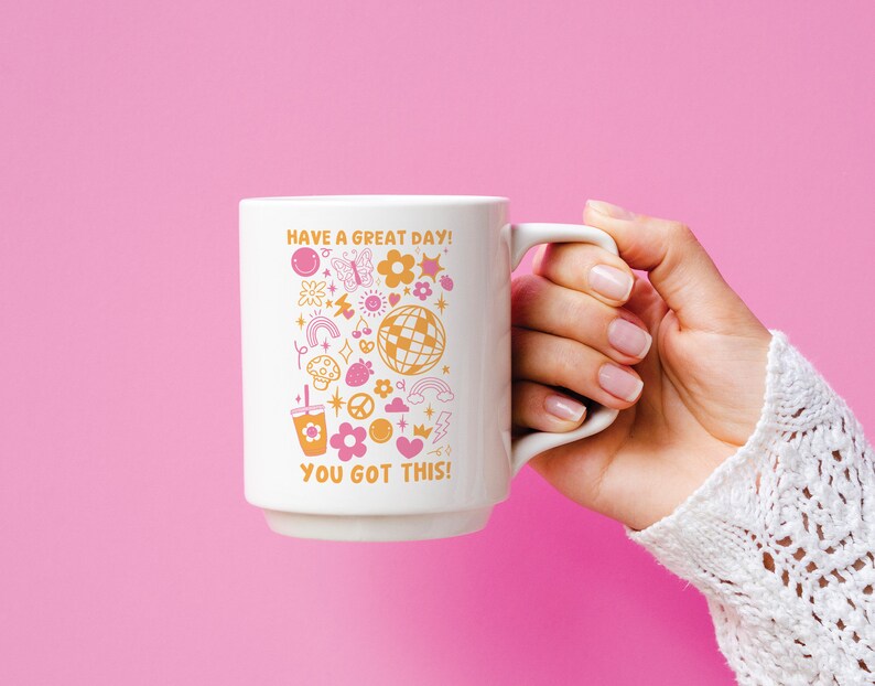 Puede incluir: Taza de cer&aacute;mica blanca con un dise&ntilde;o rosa y naranja que presenta una variedad de ilustraciones, incluyendo una mariposa, un arco&iacute;ris, un signo de paz, un coraz&oacute;n, una bola de discoteca y una cereza. El texto en la taza dice "Have a great day! You got this!"