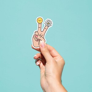 May include: A sticker of a hand making the peace sign with a smiley face on one finger and a daisy on the other finger.