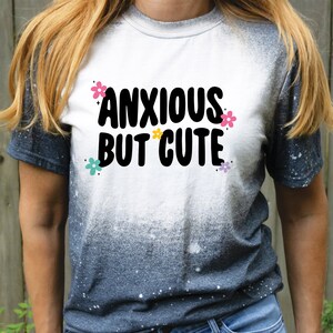 May include: A white t-shirt with a black and white graphic that says "Anxious But Cute" with colourful flowers around the text. The shirt is a tie-dye style with a white to black gradient.