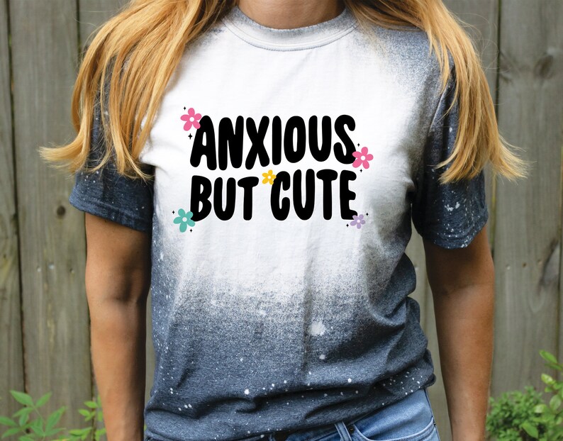 May include: A white t-shirt with a black and white graphic that says "Anxious But Cute" with colourful flowers around the text. The shirt is a tie-dye style with a white to black gradient.