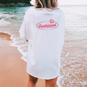May include: A white t-shirt with a pink oval graphic that says "Sunkissed" with a pink hibiscus flower inside the oval. The person wearing the shirt is standing on a beach.