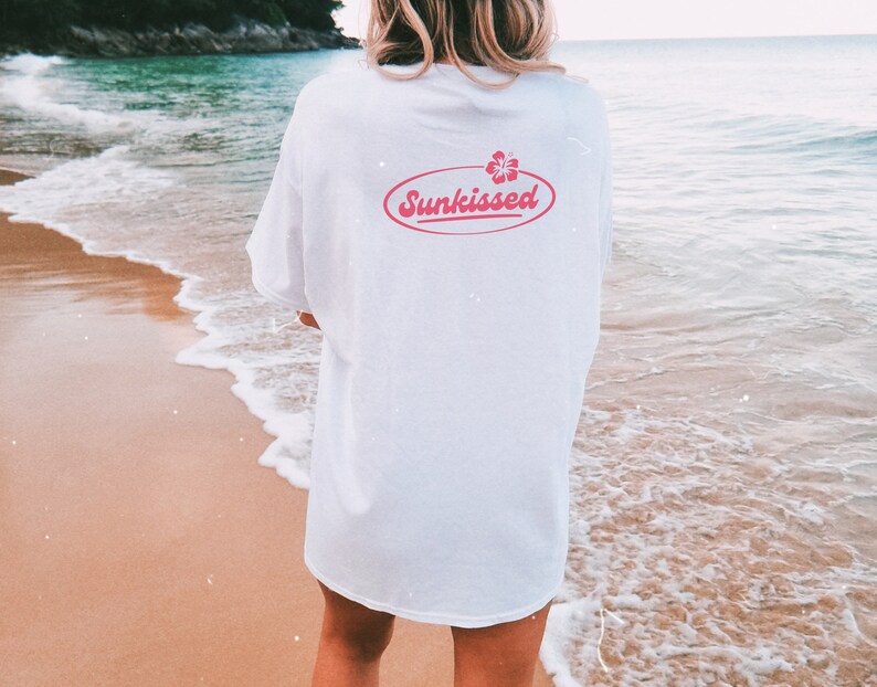 May include: A white t-shirt with a pink oval graphic that says "Sunkissed" with a pink hibiscus flower inside the oval. The person wearing the shirt is standing on a beach.