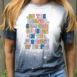 May include: A white t-shirt with a tie-dye effect on the sleeves. The shirt features a colorful graphic with the text "Be the reason someone believes in the goodness of people".
