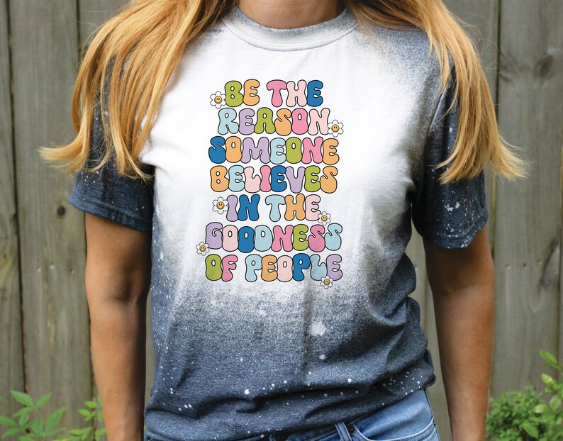May include: A white t-shirt with a tie-dye effect on the sleeves. The shirt features a colorful graphic with the text "Be the reason someone believes in the goodness of people".