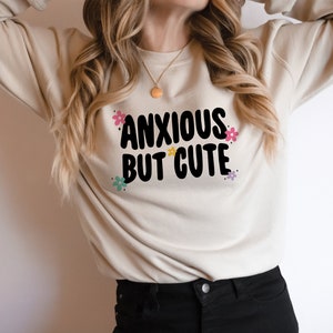 May include: A beige sweatshirt with the text "ANXIOUS BUT CUTE" in black lettering with colourful flowers surrounding the text.