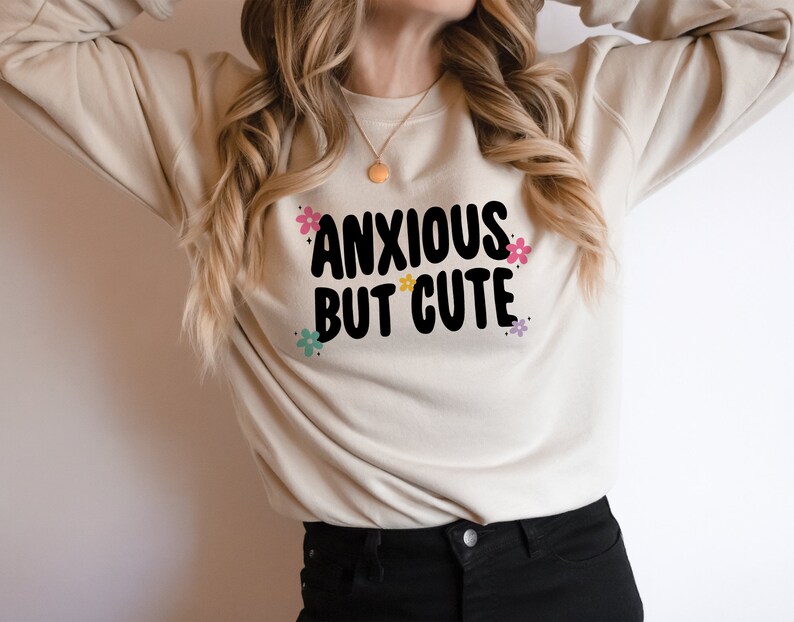 May include: A beige sweatshirt with the text "ANXIOUS BUT CUTE" in black lettering with colourful flowers surrounding the text.