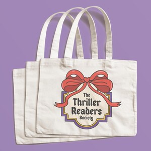 May include: Three white canvas tote bags with a red bow and ribbon design. The design includes the text "The Thriller Readers Society" in a vintage style. The bags have white handles and are set against a purple background.