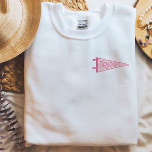 May include: White sweatshirt with a pink pennant design that says "Tired AF".
