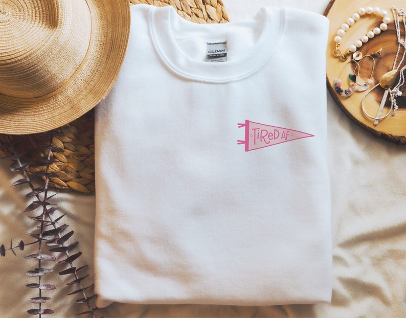 May include: White sweatshirt with a pink pennant design that says "Tired AF".