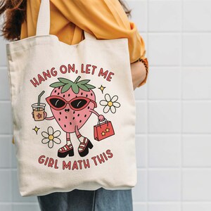 May include: A white canvas tote bag with a cartoon strawberry wearing sunglasses and holding a shopping bag. The text on the bag reads "Hang on, let me girl math this".