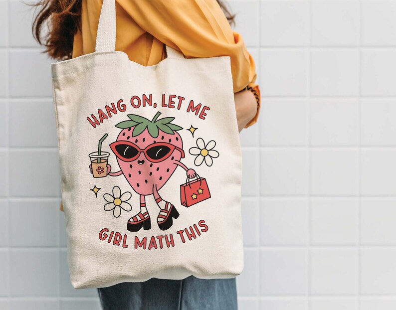 May include: A white canvas tote bag with a cartoon strawberry wearing sunglasses and holding a shopping bag. The text on the bag reads "Hang on, let me girl math this".