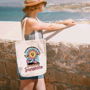 Op de afbeelding: Een witte canvas tote bag met een kleurrijke afbeelding van een lachende zon met de tekst "I'm a happy go lucky ray of freaking sunshine".