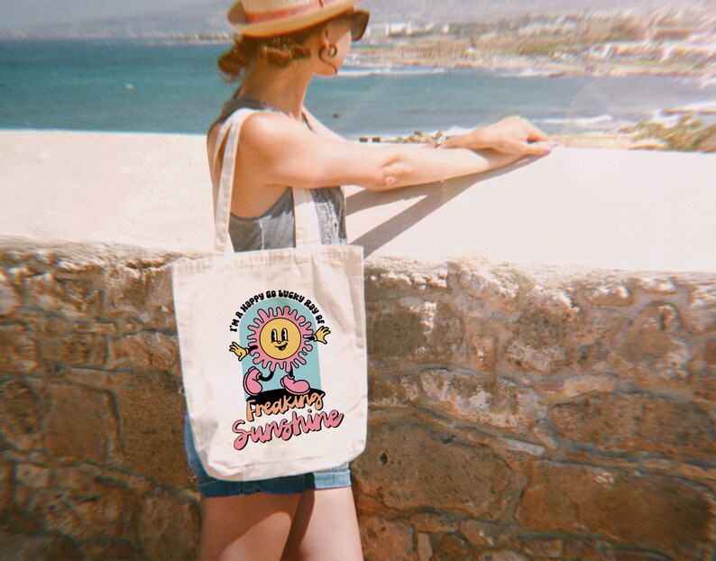 Op de afbeelding: Een witte canvas tote bag met een kleurrijke afbeelding van een lachende zon met de tekst "I'm a happy go lucky ray of freaking sunshine".