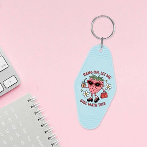 May include: A light blue key chain shaped like a motel key with a cartoon strawberry wearing sunglasses and holding a drink and a purse. The text on the key chain reads "Hang on, let me girl math this".